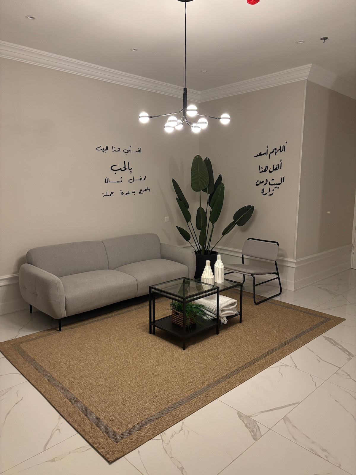 A light gray sofa is positioned in a modern living area, accompanied by a minimalistic black chair and a glass coffee table. A large potted plant adds a touch of greenery, while Arabic calligraphy is displayed on the beige walls, enhancing the space's character.