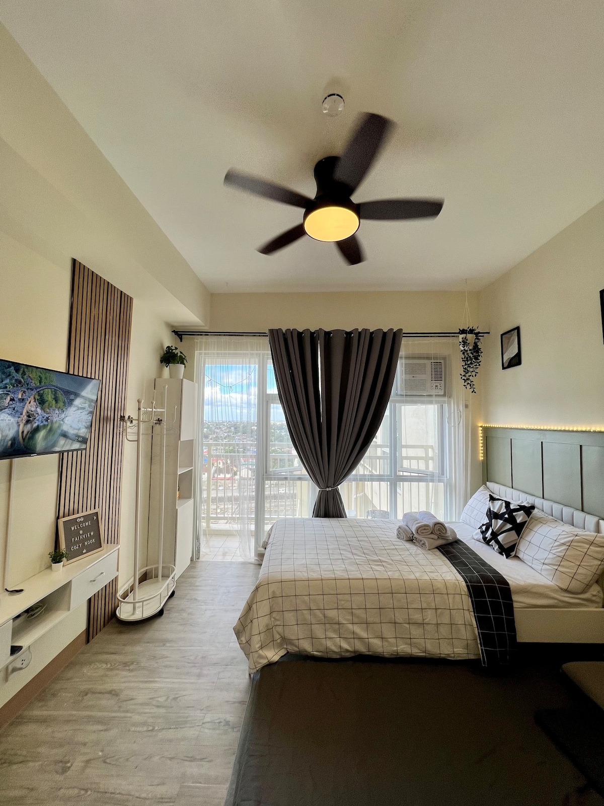 A well-lit studio room features a spacious bed with checkered bedding and decorative throw pillows. A ceiling fan circulates air, while large windows provide a view of the city. Blackout curtains frame the window, and a television is positioned on the wall opposite the bed.