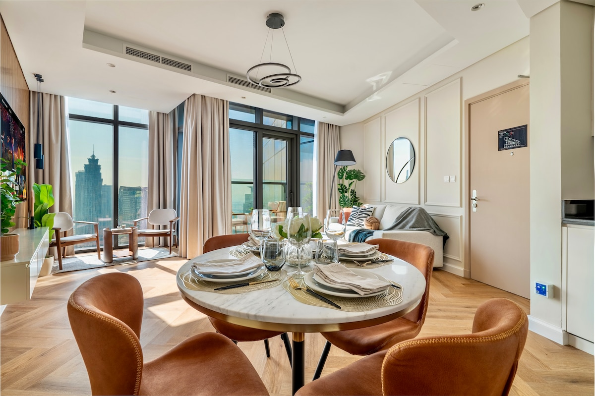A bright living area features a round dining table set for four, surrounded by elegant leather chairs. In the background, expansive windows reveal stunning city views. A cozy lounge space is arranged with a modern sofa and stylish floor lamp, complemented by warm wood accents.