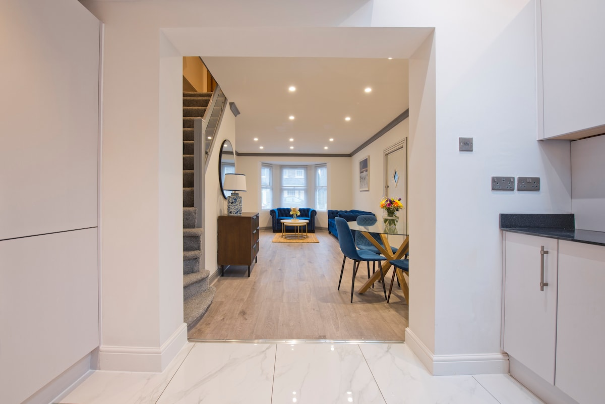 An open doorway frames a spacious living area, featuring modern furnishings and natural light. A staircase leads to the upper level, while a dining set with blue chairs is positioned next to a decorative side table. Soft colors create a harmonious ambiance throughout the space.