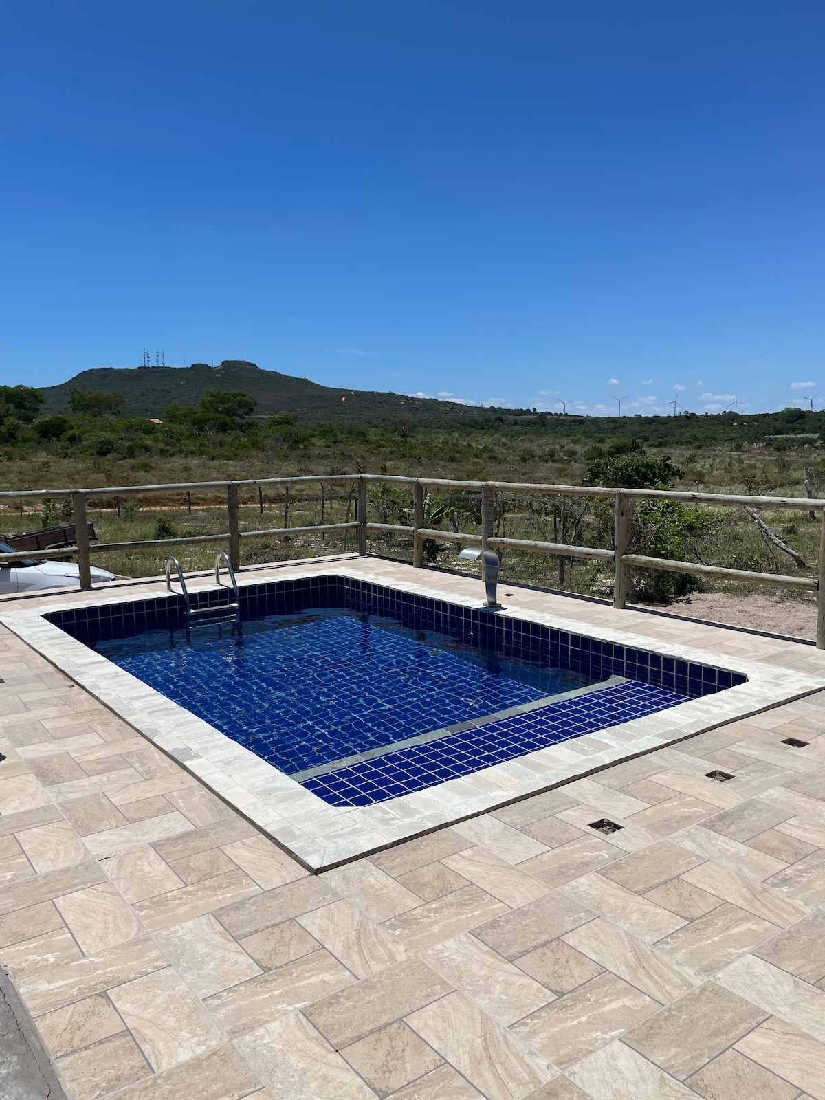A private swimming pool with blue tiles is surrounded by a stone deck. A wooden railing overlooks the natural landscape and distant hills under a clear blue sky, creating a serene outdoor space for relaxation.
