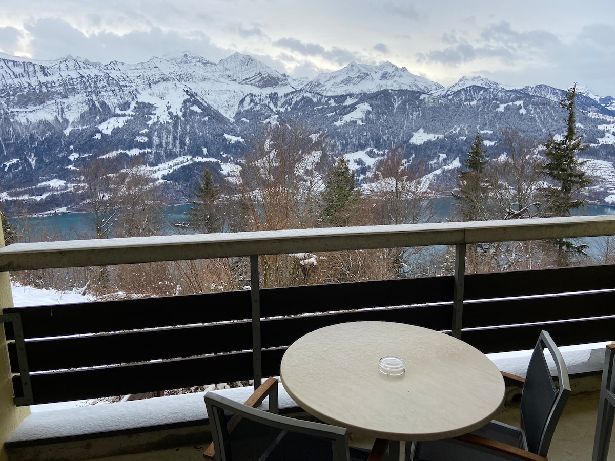 A spacious balcony showcases stunning views of snow-capped mountains and the Thunersee. A round table with chairs is positioned on the balcony, inviting relaxation while taking in the scenic landscape.