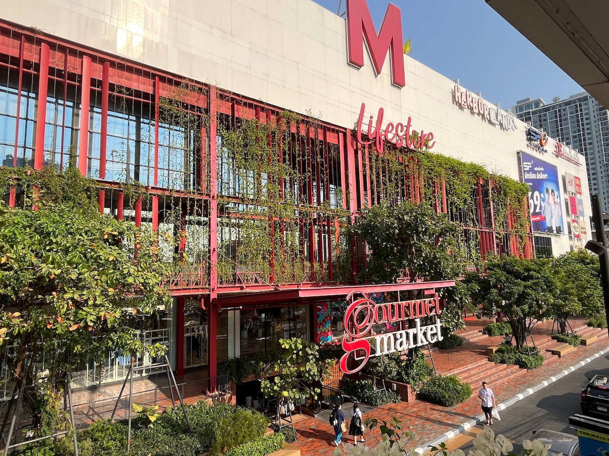 An urban shopping center is featured, characterized by a modern facade with vibrant greenery intertwined throughout. The entrance is designated by a prominent sign reading 'Gourmet Market.' Pedestrians can be seen walking along the paved pathway, contributing to the lively atmosphere of the area.