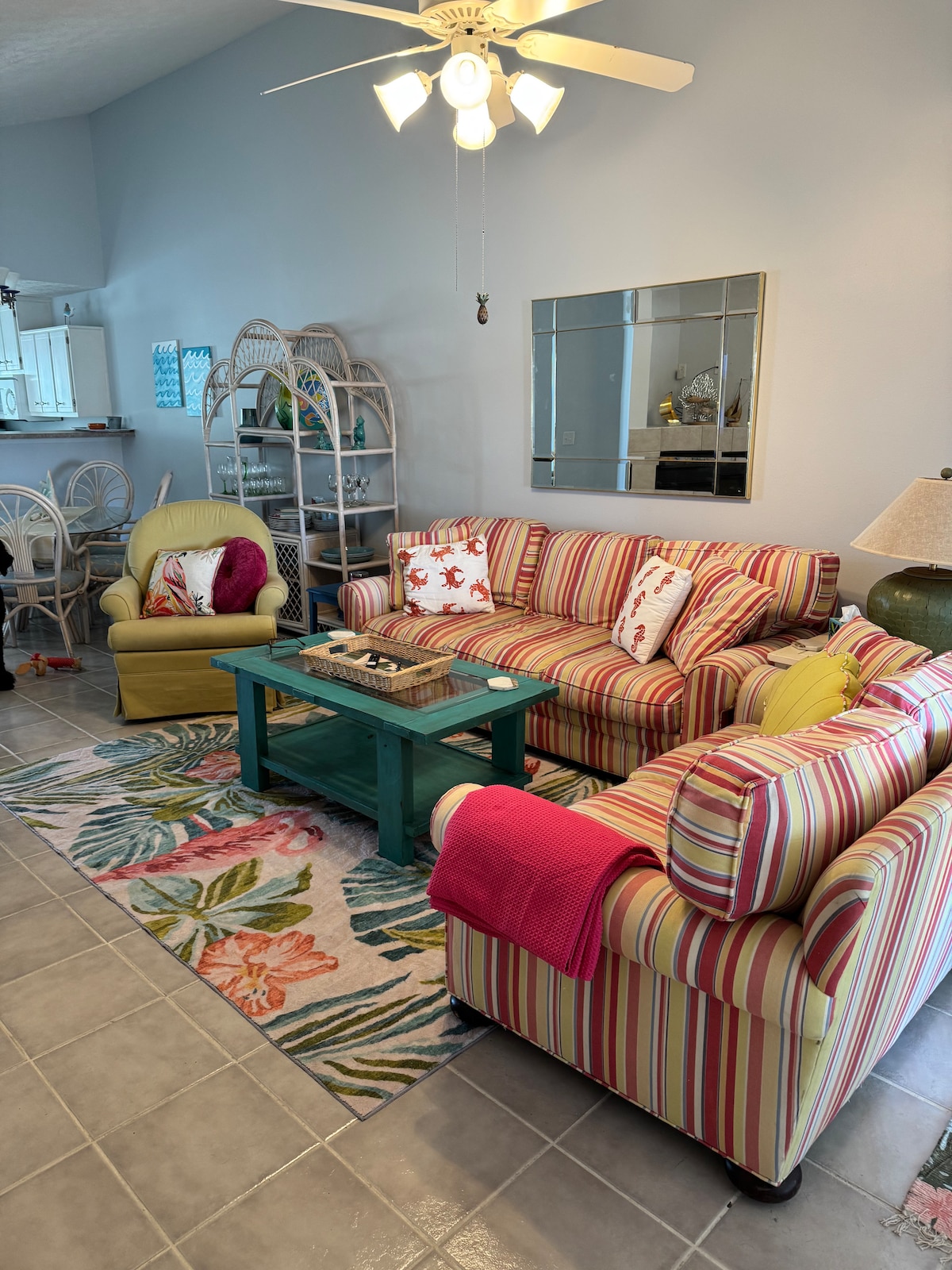 The living area features a striped, spacious sofa adorned with decorative pillows, complemented by a cheerful armchair. A teal coffee table is centered on a vibrant floral rug, and a ceiling fan provides airflow. Light reflects off a wall mirror, enhancing the welcoming ambiance.