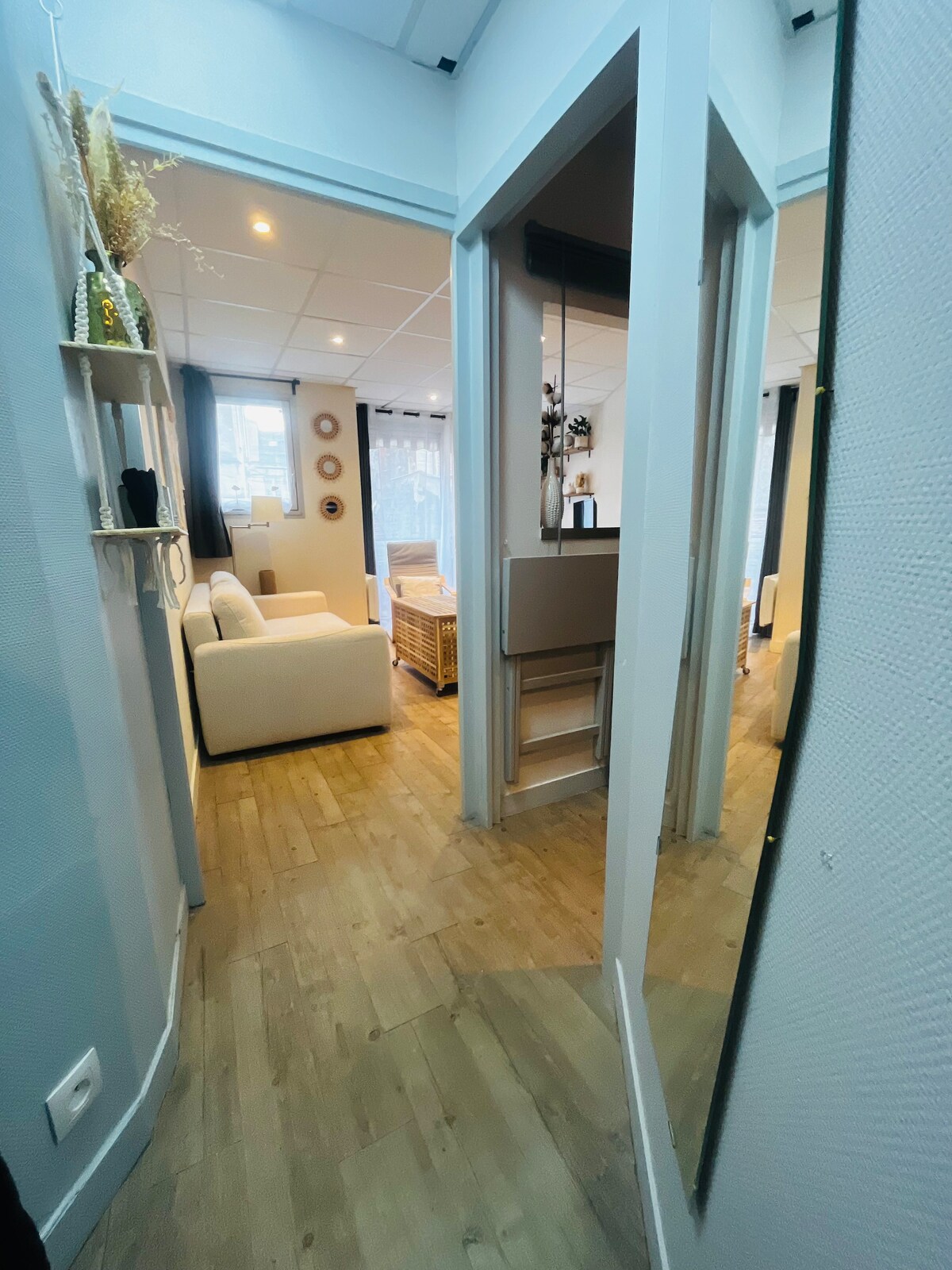 The image displays a narrow hallway leading into a bright living space. A large mirror reflects the light from the room, which features a comfortably arranged white sofa and wooden furniture. Natural light filters through nearby windows, creating an inviting atmosphere.