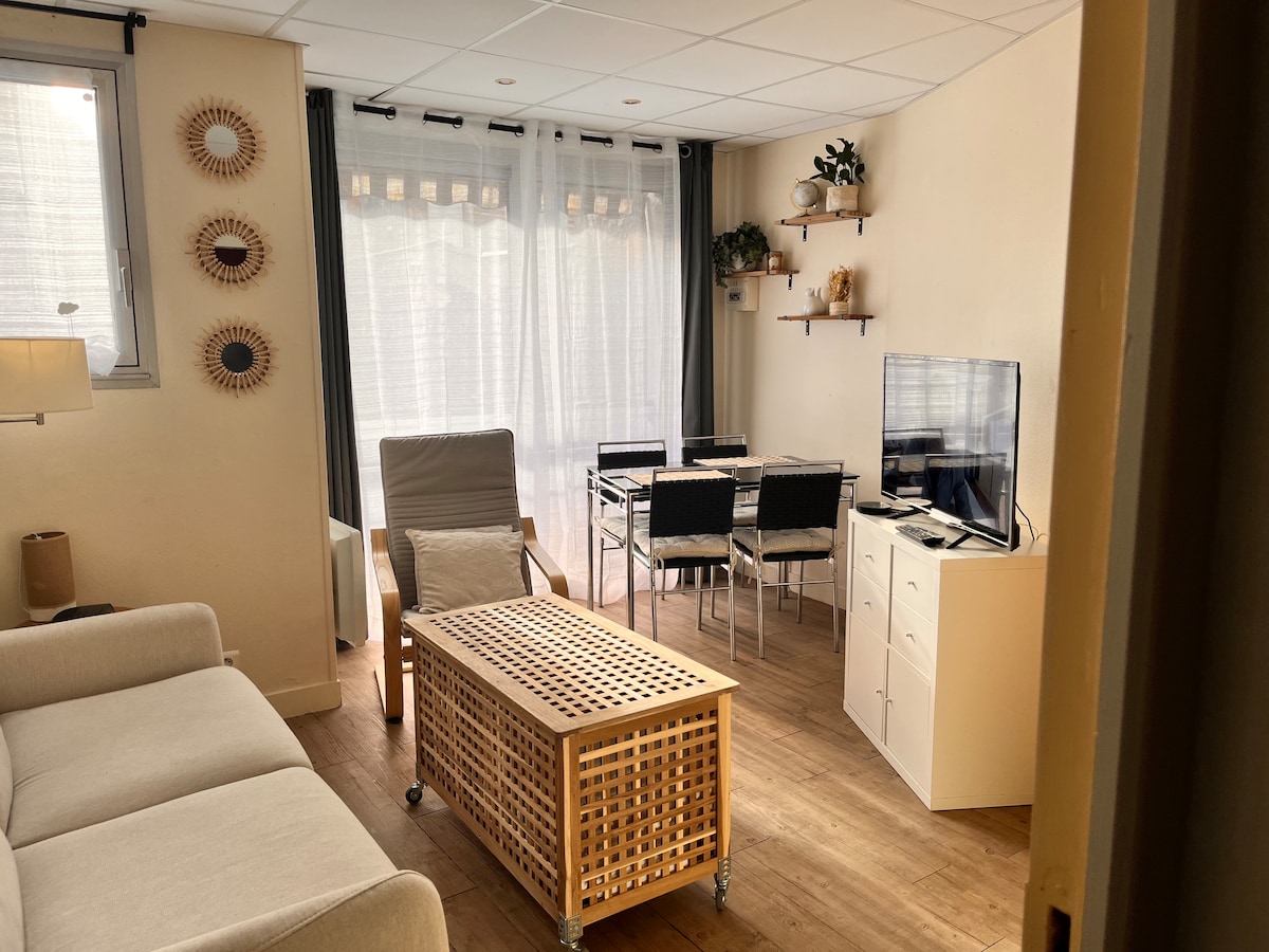 A bright living area features a comfortable sofa and a stylish coffee table with a woven design. A television is positioned for viewing, and a dining table with several chairs is visible nearby. Sheer curtains allow natural light to fill the space, creating a welcoming atmosphere.