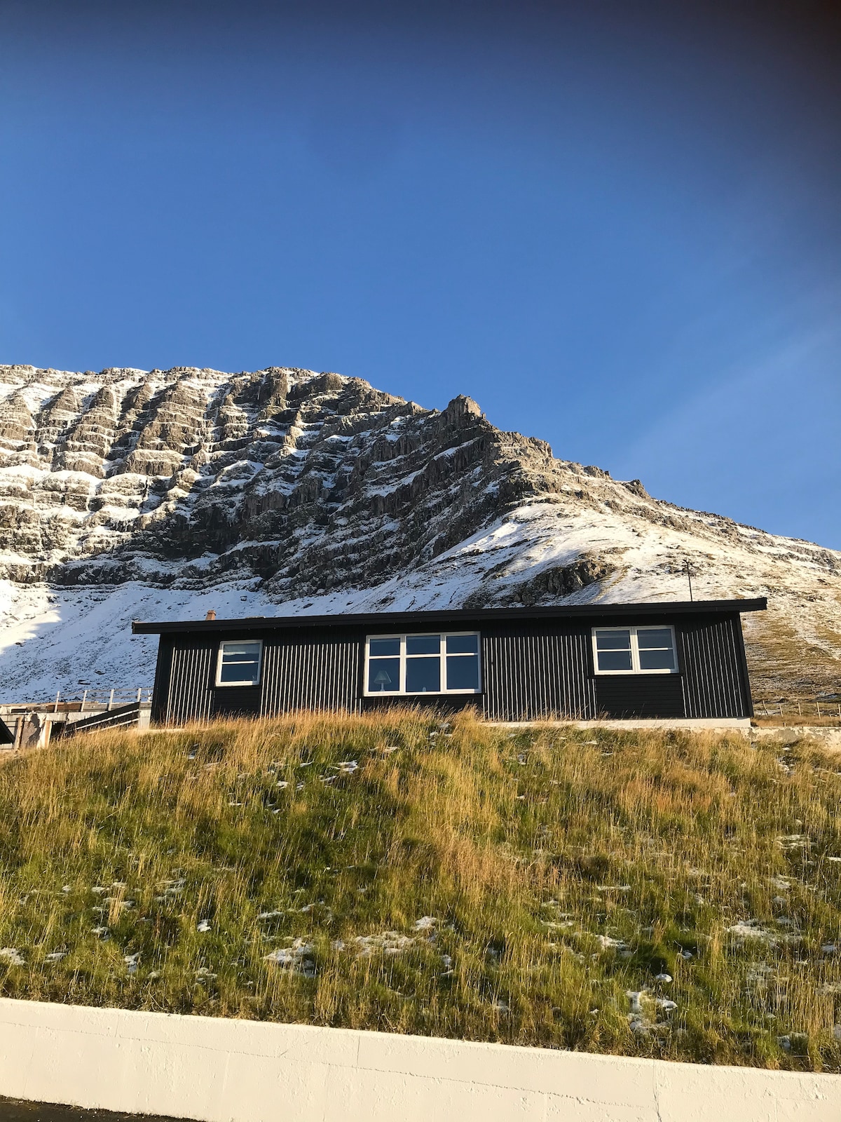 A traditional black house is situated on a grassy slope, framed by a mountainous backdrop. The clear blue sky creates a striking contrast with the snow-capped peaks, while expansive windows reflect the natural surroundings.