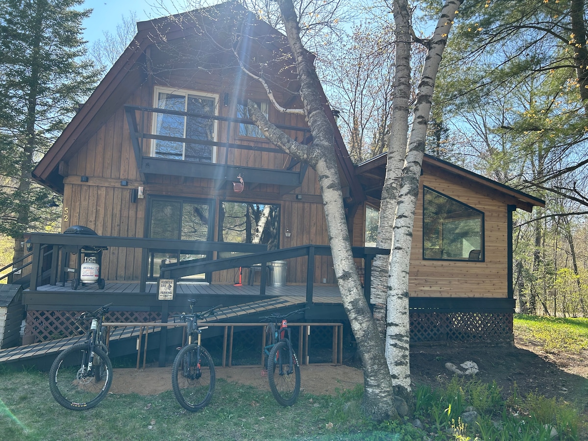 A charming chalet is set among trees, featuring a sloped wooden ramp leading to a spacious deck. Two bicycles are parked in front, with large windows providing natural light and views of the surrounding landscape. The structure has a combination of wood and glass elements.