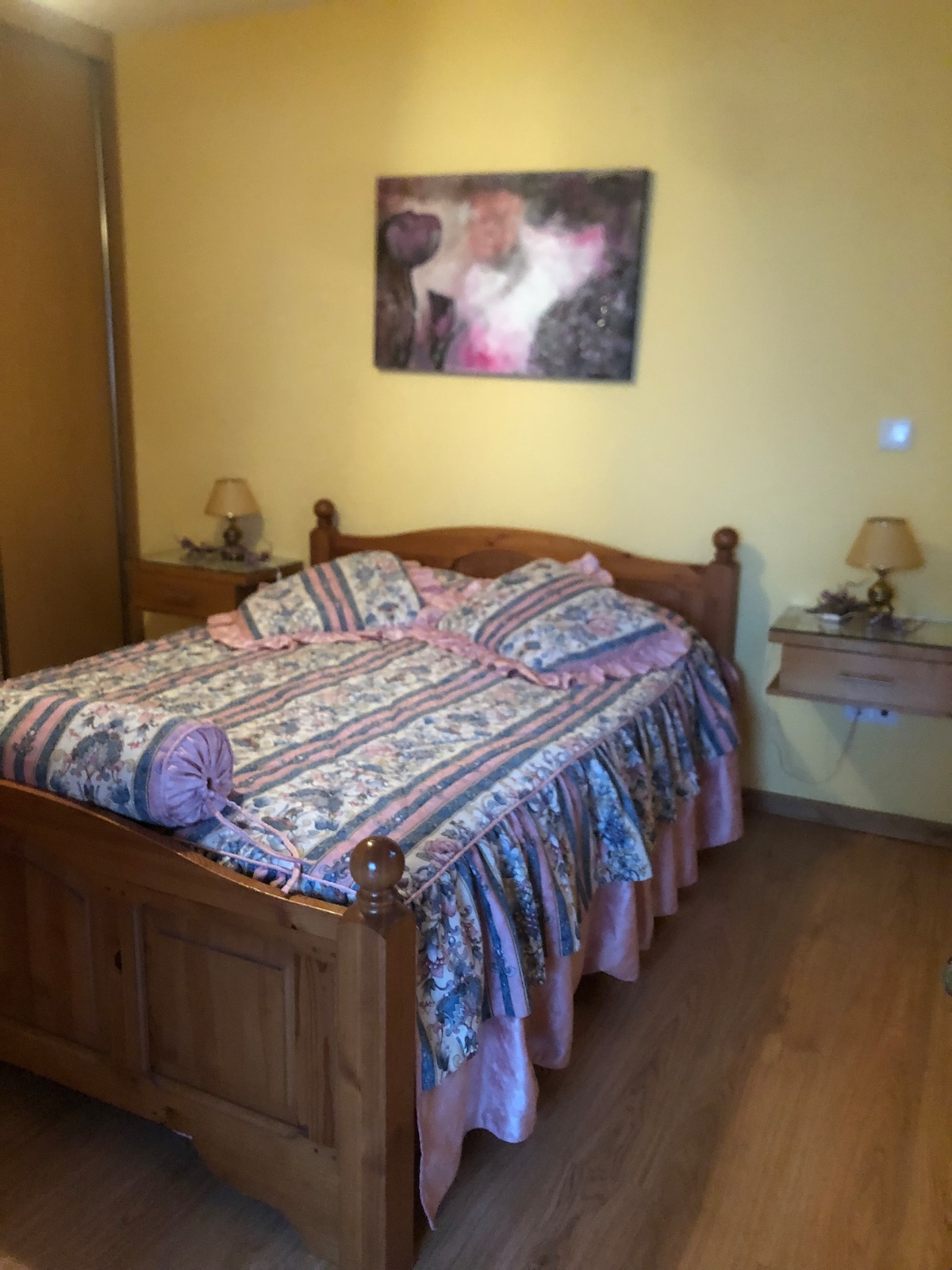 A cozy sleeping area features a wooden bed with a patterned bedspread and decorative pillows. Soft bedside lamps are positioned on either side of the bed, while a colorful wall art piece adds a touch of character to the warm yellow walls.