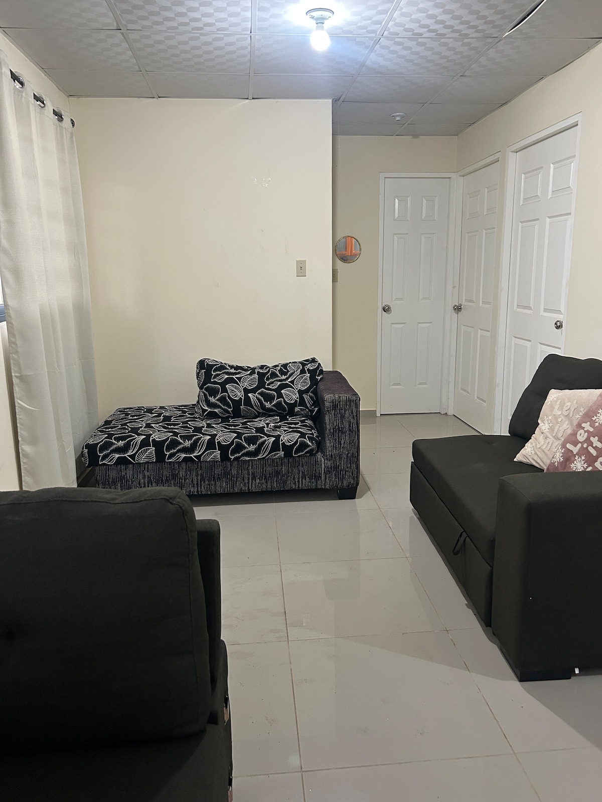 A living area showcases a dark-colored sofa and a chaise lounge adorned with a floral pattern. Light-colored walls are complemented by two white doors, and natural light enters through a nearby window covered by sheer curtains. A ceiling light provides illumination.