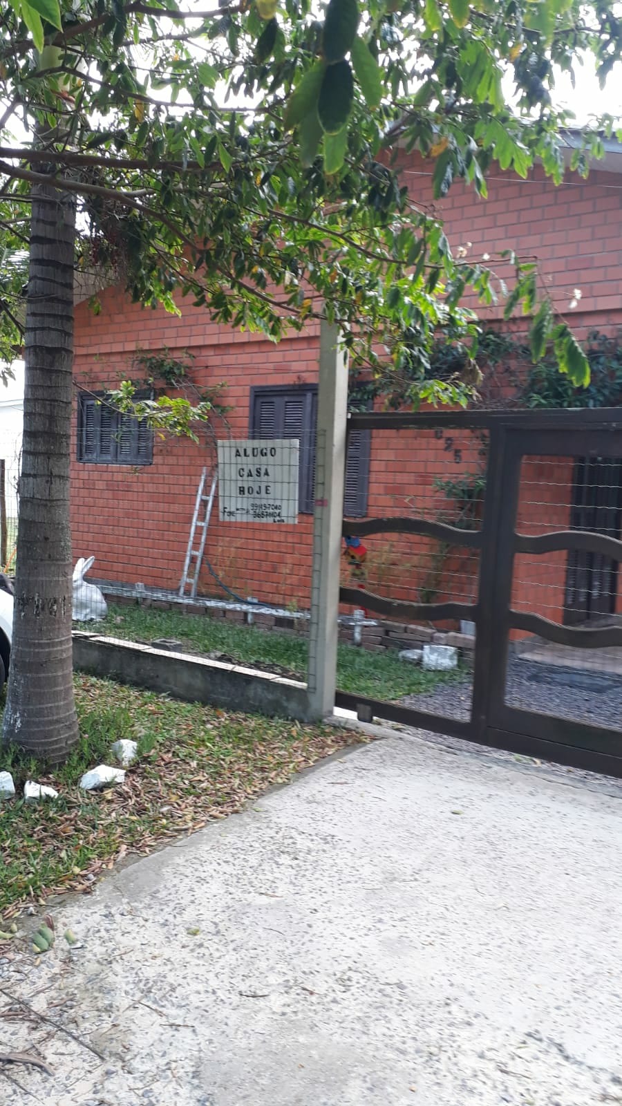 The exterior of a brick residence is visible, framed by lush greenery. A concrete driveway leads to a secure gate, with the property name displayed prominently. A ladder leans against the house, suggesting ongoing maintenance or landscaping work.