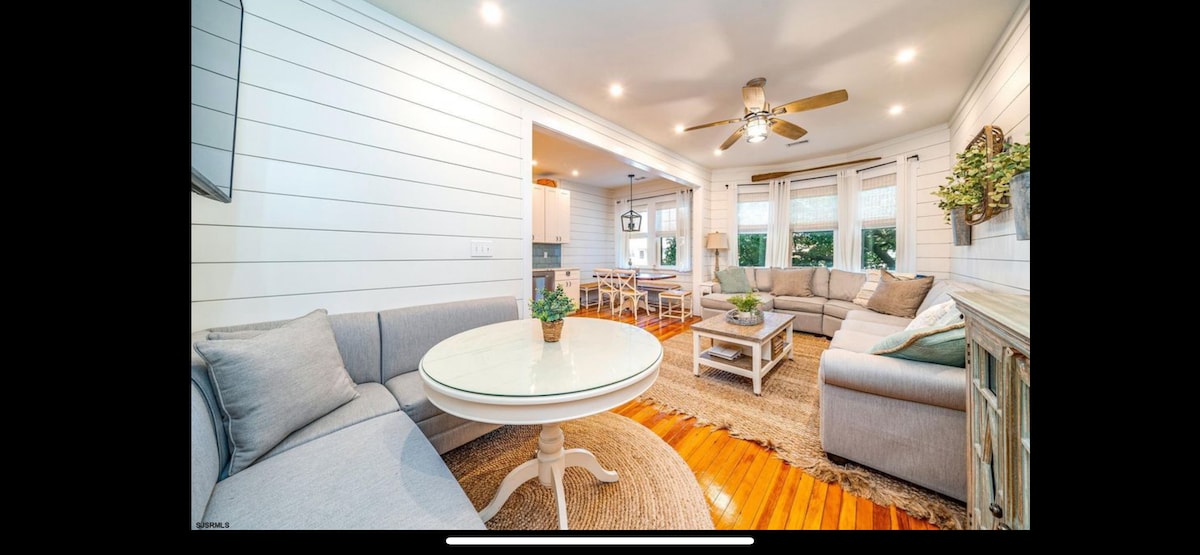 A bright living area features shiplap walls and wooden floors. A circular table is positioned in front of a sectional sofa with soft cushions. Natural light enters through multiple windows, while a ceiling fan adds comfort to the space. Decorative plants are placed thoughtfully.