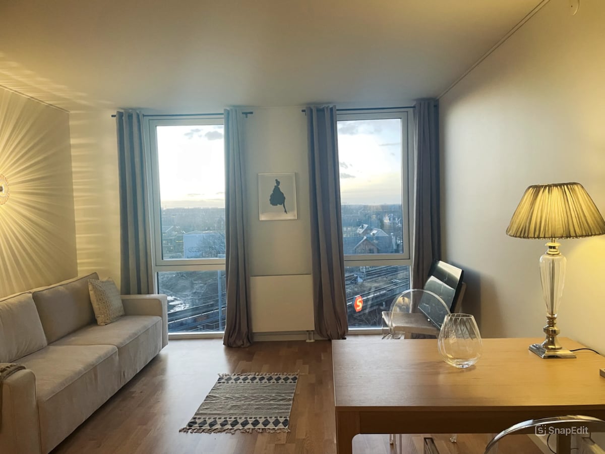 A modern living space is presented with a light-colored sofa positioned near two large windows offering expansive city views. A wooden table sits nearby, accompanied by a clear vase, and a comfortable chair is located adjacent to the table, with a stylish lamp providing warm illumination.