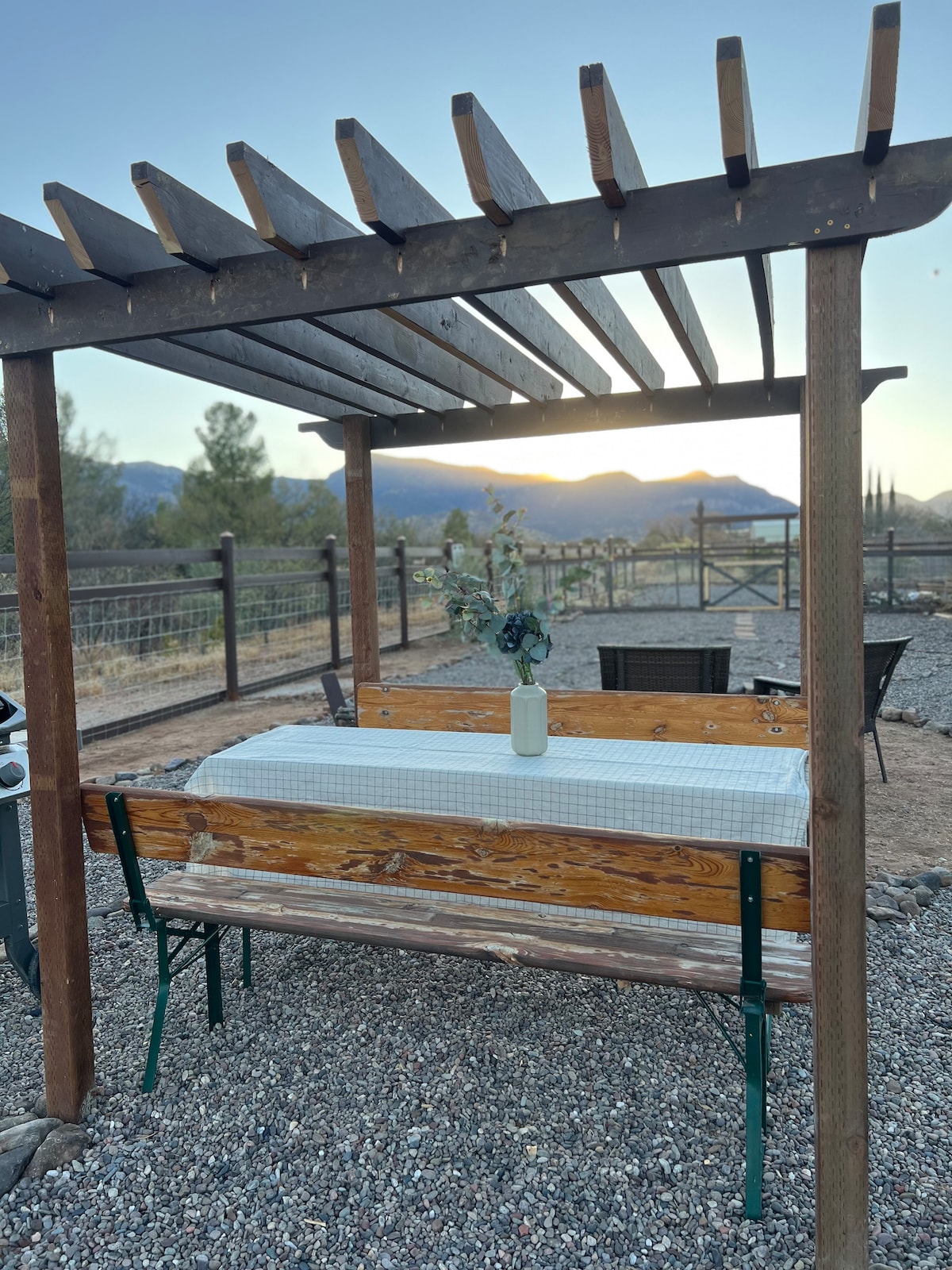 An outdoor dining area features a wooden table with a vase of dried flowers, shaded by a pergola. Surrounding the table are metal chairs, with gravel underfoot. The backdrop reveals distant mountains, softly illuminated by the setting sun.