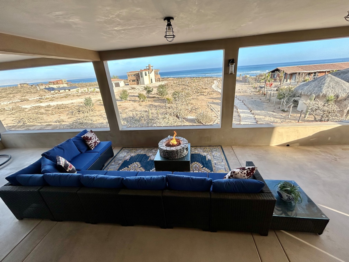 A spacious outdoor seating area is showcased, featuring a large blue sectional sofa arranged around a circular fire pit. The area overlooks a sandy landscape with distant ocean views, providing a comfortable spot for relaxation while enjoying the coastal scenery.