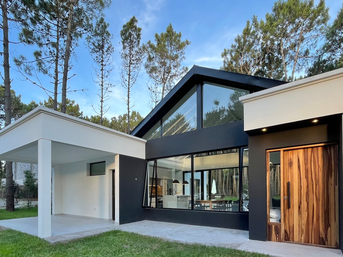 A modern house features a striking combination of black and white exteriors, with expansive windows allowing natural light to illuminate the interior spaces. Surrounding tall trees enhance the serene setting, while the front entrance showcases a wooden door with unique grain patterns.