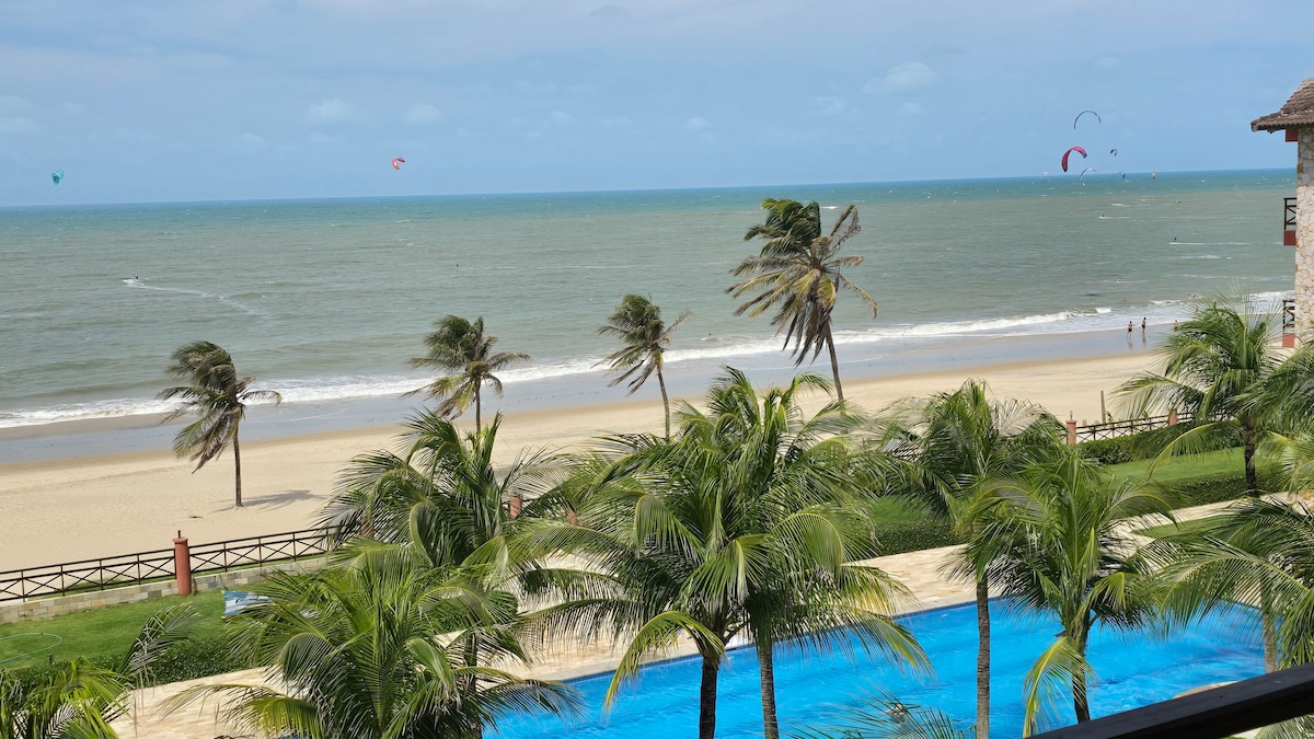 A beachfront view captures gentle waves lapping against the sandy shore, with kitesurfers gliding across the water in the distance. Lush palm trees frame the scene, while a shimmering blue pool is visible in the foreground, complementing the tranquil coastal setting.