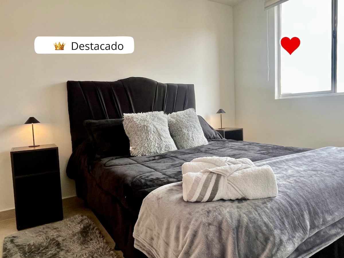 The main bedroom features a spacious bed with a dark upholstered headboard, complemented by soft throw pillows. Nightstands with lamps are positioned on either side. A large window allows natural light to fill the room, partially revealing views of the outside.
