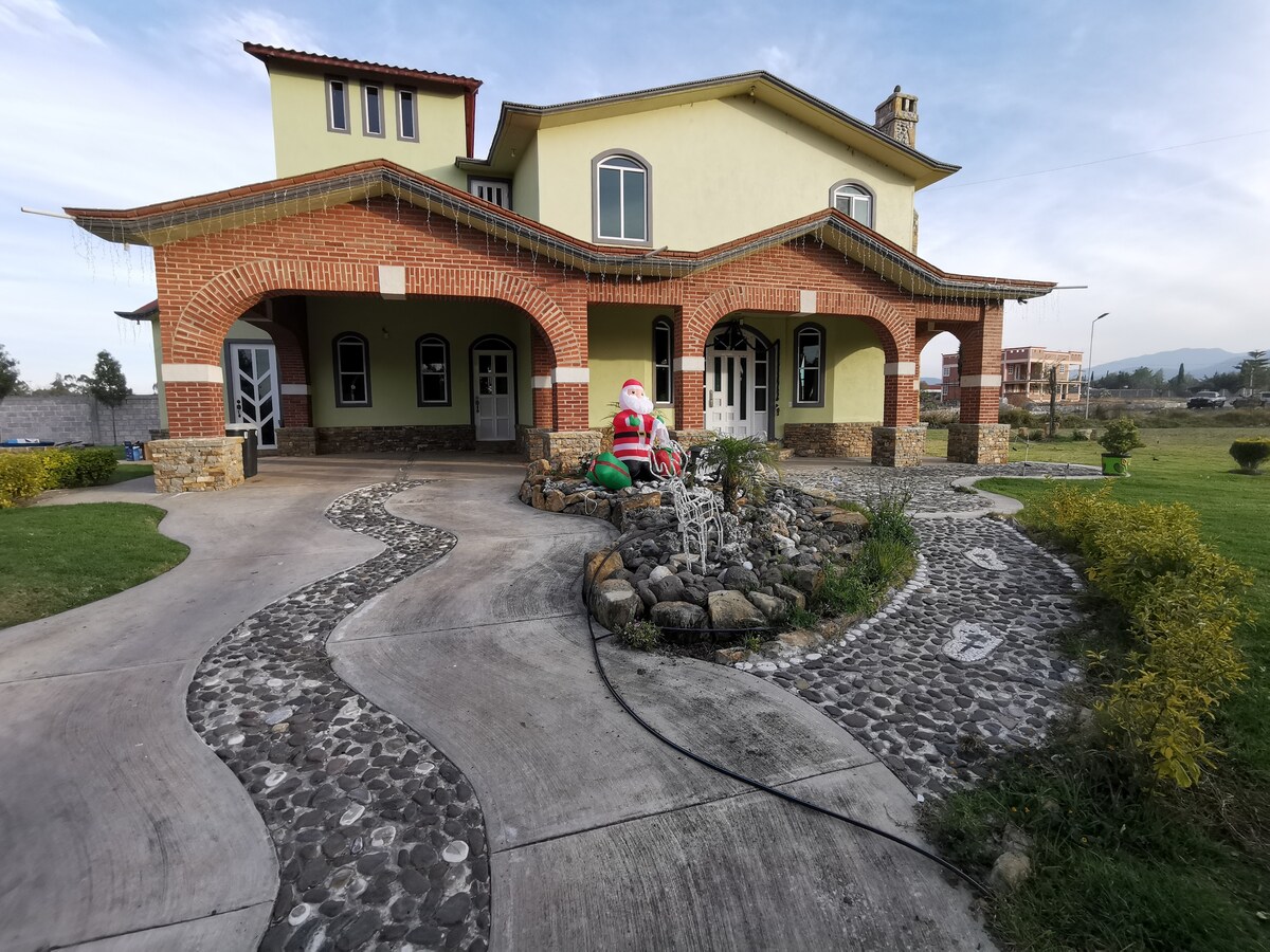 A spacious building with a light yellow exterior features an arched entrance and a pathway made of cobblestones. A decorative Santa figure is positioned among landscaped stones and green bushes, creating an inviting entryway.