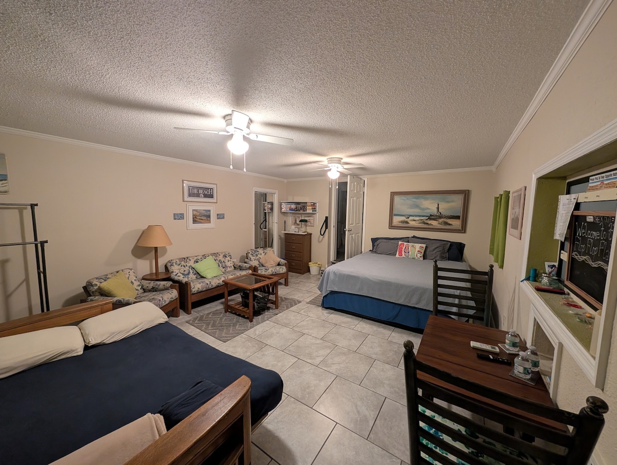 A spacious efficiency guest suite is displayed, featuring a large bed and a pull-out sofa bed. A comfortable seating area includes patterned chairs and a wooden dining table. Bright lighting from overhead fans and natural light contributes to the open atmosphere.
