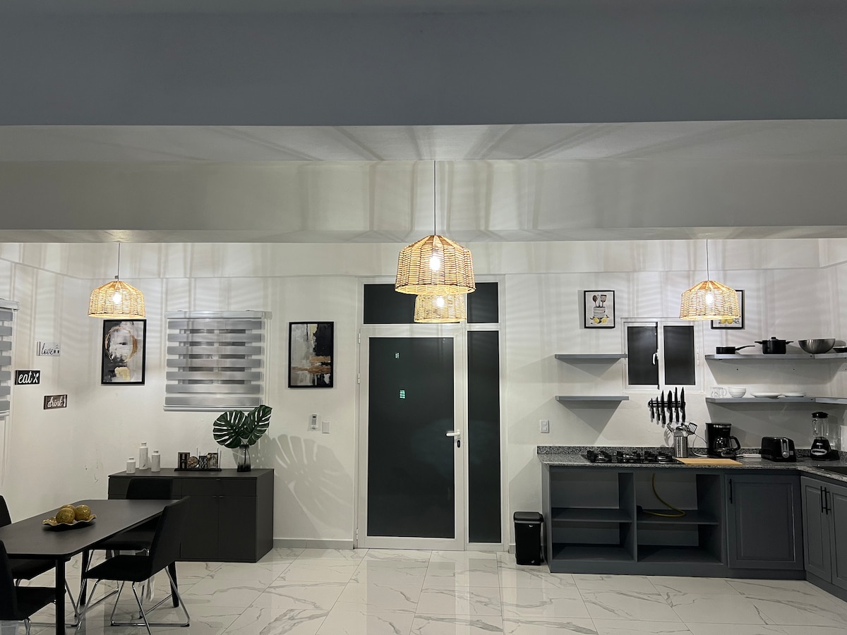 An open kitchen and dining area features modern cabinetry and sleek appliances, including a coffee machine and kitchen utensils. Three pendant lights hang above the dining table, which is surrounded by black chairs. Light-colored tiles reflect natural light, enhancing a spacious atmosphere.