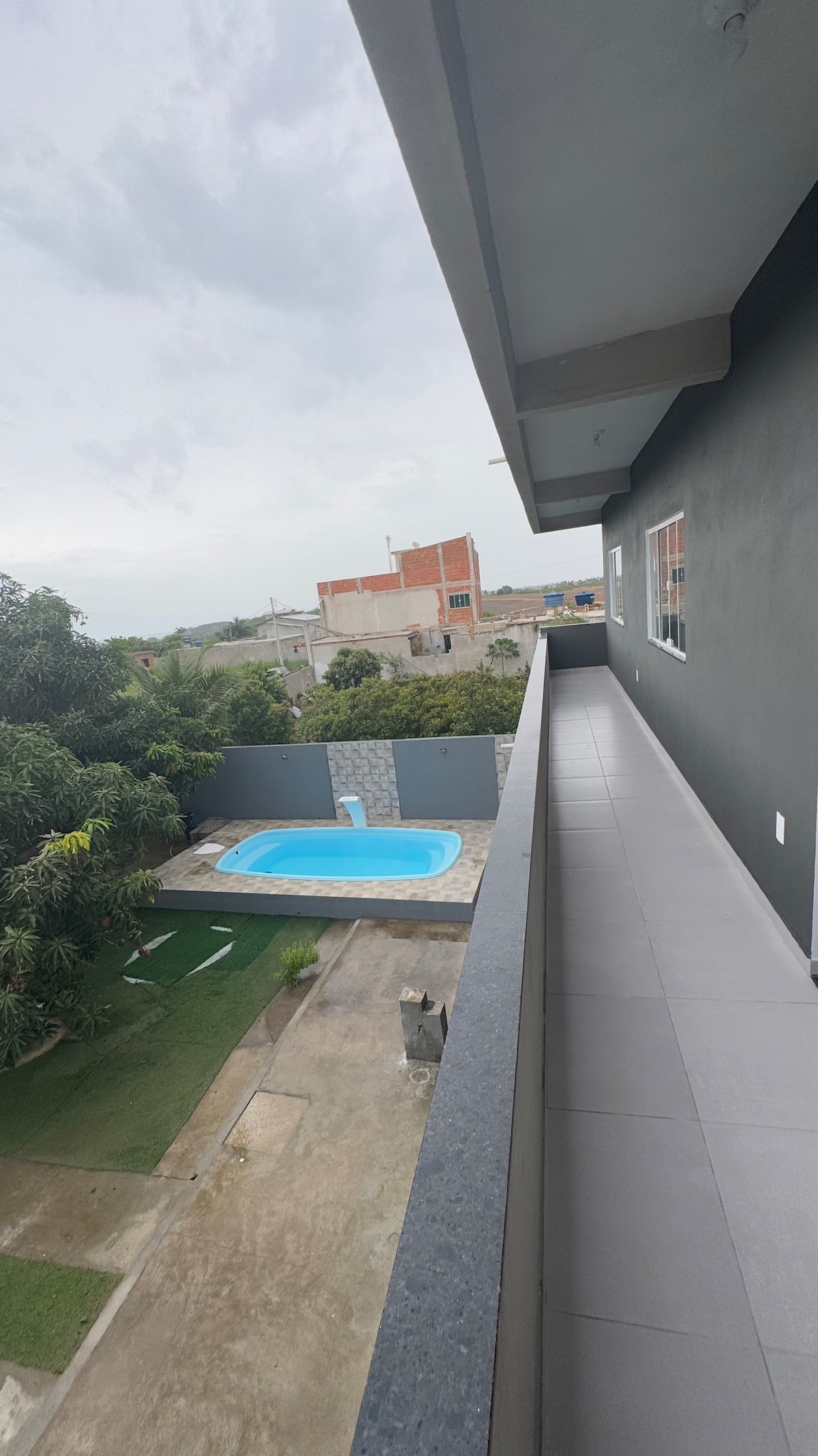 A wide balcony offers a view of a private pool on a stone patio, surrounded by greenery. The scene includes well-maintained grass and a glimpse of nearby structures. Soft natural light enhances the spacious outdoor area, characterized by a clean, modern design.
