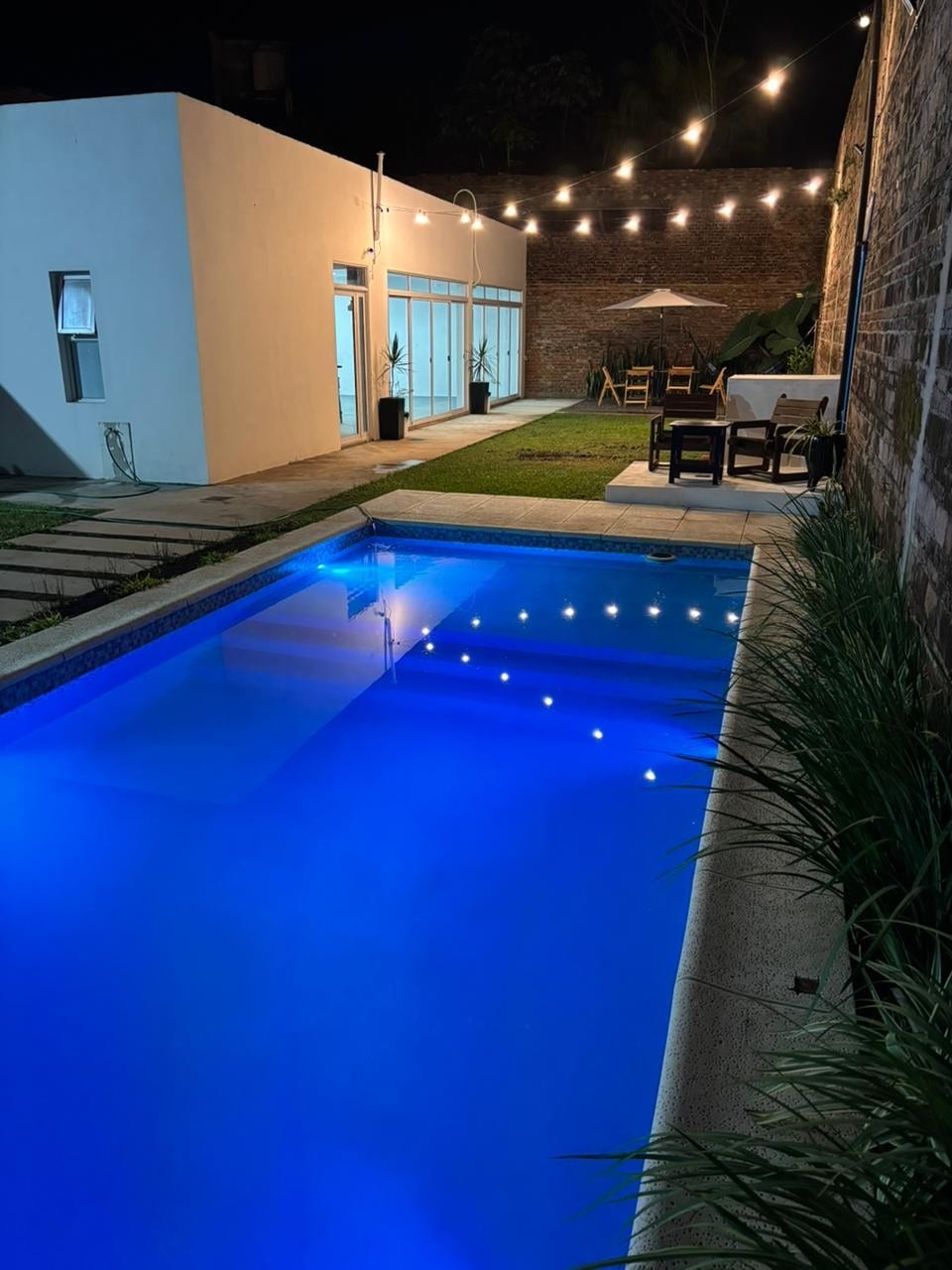A tranquil outdoor space features a well-lit swimming pool with illuminated blue water. A wooden dining area is situated nearby, under soft string lights, enhancing the inviting atmosphere. A grassy area complements the scene, while modern glass doors offer access to the interior.