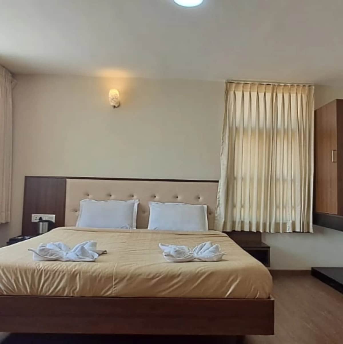 A spacious bedroom features a soft beige bedspread atop a wooden platform bed. Two plush pillows are arranged on the headboard, with neatly folded white towels placed at the foot of the bed. Natural light filters through a large window with cream-colored curtains.