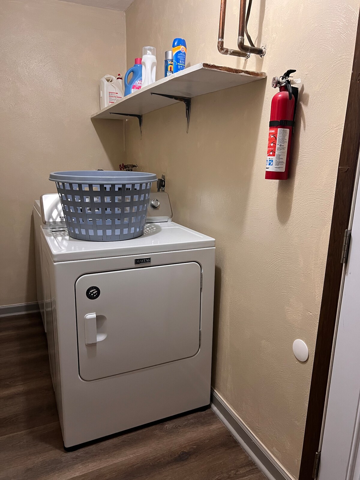 A laundry area is equipped with a white dryer and a laundry basket filled with clothes. Above the dryer, shelving is mounted on the wall, holding various laundry supplies. A fire extinguisher is securely attached to the wall nearby.