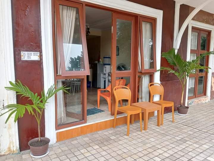 An outdoor seating area features two orange chairs and a small table positioned in front of large glass doors. Potted plants add greenery, while the interior reveals glimpses of a kitchen space beyond.