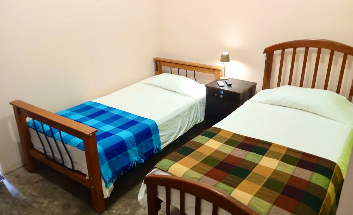 A simple sleeping area is furnished with two single beds, each dressed in light linens. One bed features a blue plaid throw, while the other has a green plaid blanket. A bedside table with a lamp sits between the beds, providing a cozy ambiance.