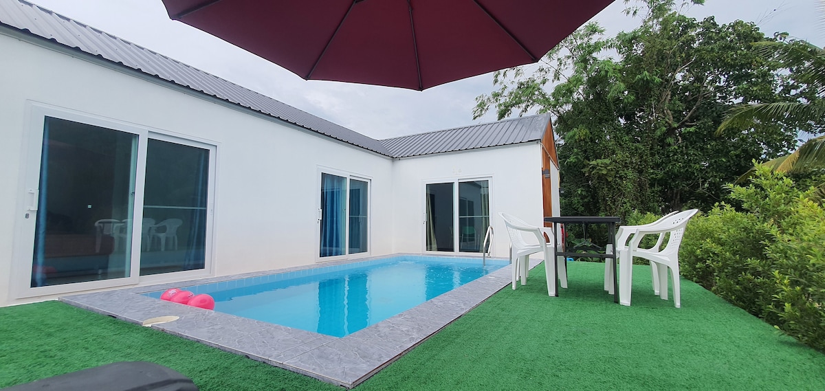 A modern outdoor area features a private pool surrounded by green grass. White lounge chairs are positioned next to the pool. An umbrella provides shade over a small table, enhancing the functionality of the space. Lush greenery borders the area, contributing to the serene environment.