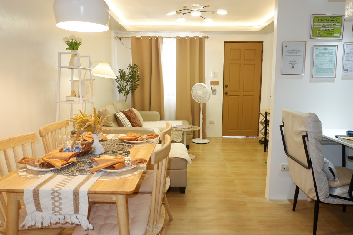 A dining area features a wooden table set for four, complemented by natural woven accents. Soft lighting illuminates a cozy living space equipped with a sofa and an oscillating fan. Light brown curtains frame the window, allowing natural light to fill the room.
