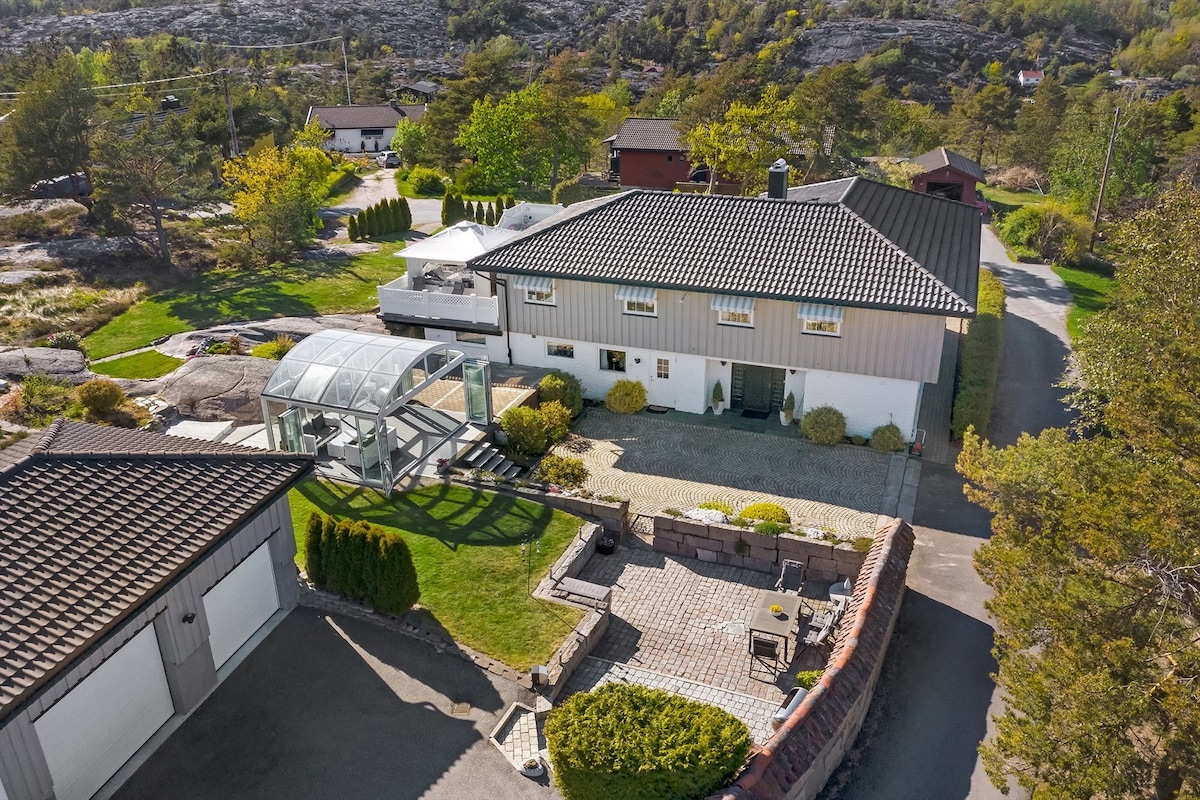 An aerial view captures a spacious property featuring a well-maintained garden with green lawn, stone pathways, and an outdoor seating area. A covered patio is seen adjacent to the house, which has a mixture of light and dark exterior walls. The surrounding area includes trees and neighboring buildings.