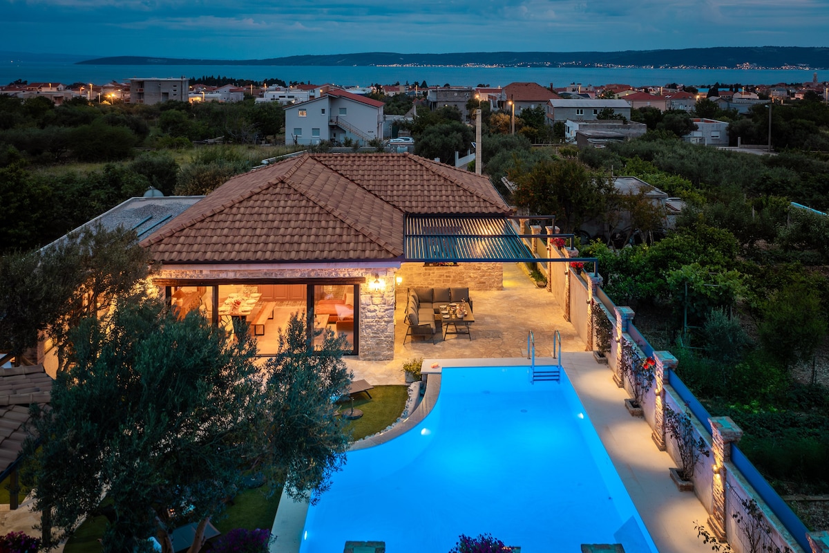 An aerial view showcases the villa with a warm glow emanating from the windows. The 55 square meter pool reflects twilight hues, surrounded by lush greenery. The outdoor dining area is visible, complemented by the terrace leading into the villa, creating a serene atmosphere against the dusk sky.