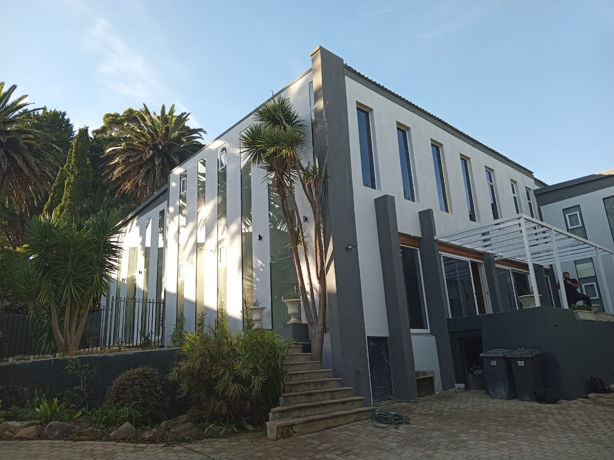 A modern building features a clean exterior with large windows that allow natural light to flow inside. Palm trees and lush greenery surround the entrance, where steps lead up to a covered patio area, creating an inviting atmosphere that blends with the garden setting.
