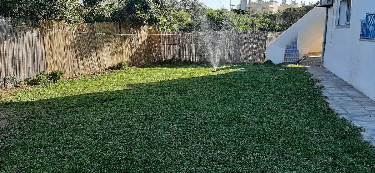 A spacious lawn is surrounded by a natural wooden fence, featuring green grass and an automatic sprinkler in the center. Sunlight creates distinct areas of shade and brightness, with a path leading to a building in the background.