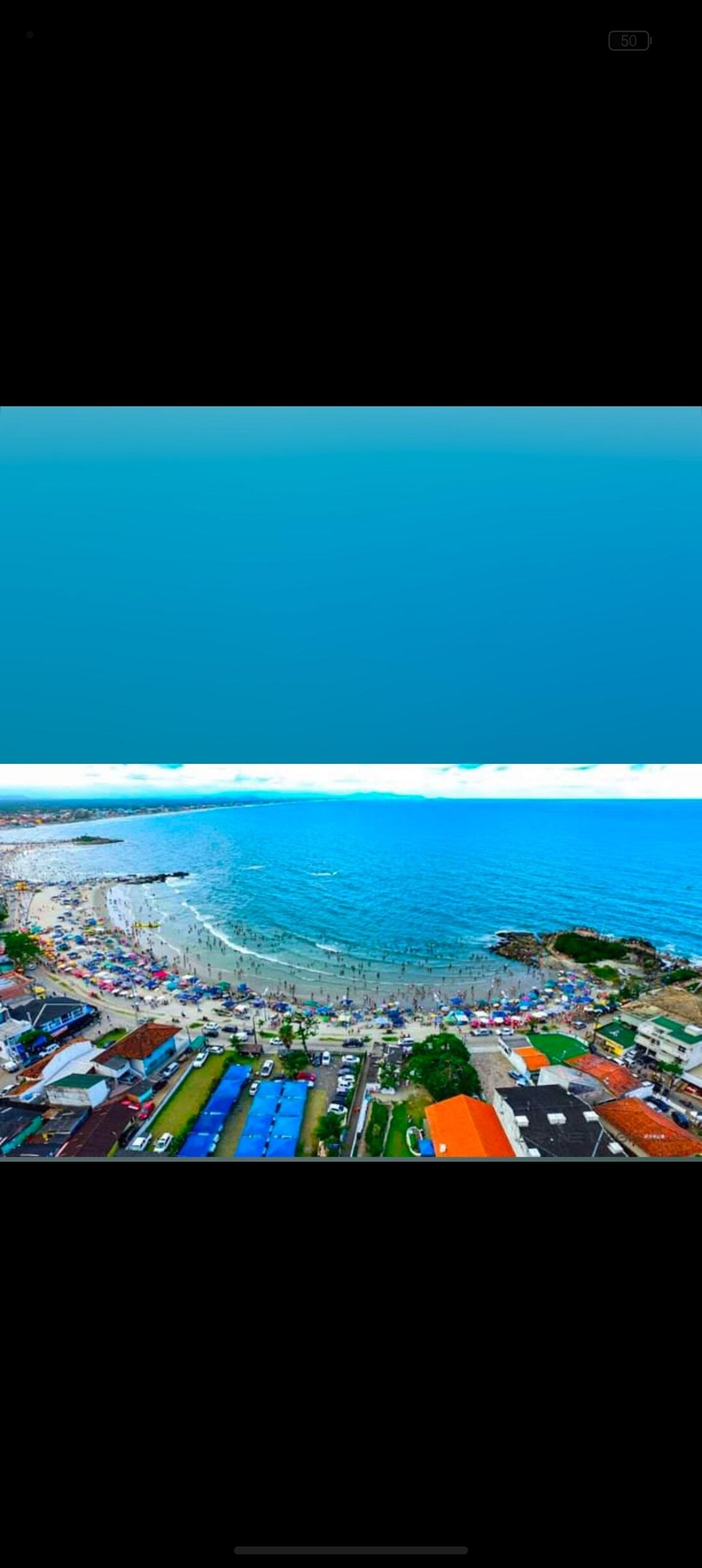 An aerial view captures the coastline and beach, showcasing a vibrant mixture of colorful umbrellas and beachgoers. The ocean displays clear blue waters, while the shoreline reveals a blend of sandy areas and picturesque waves. Nearby buildings and greenery are also visible.