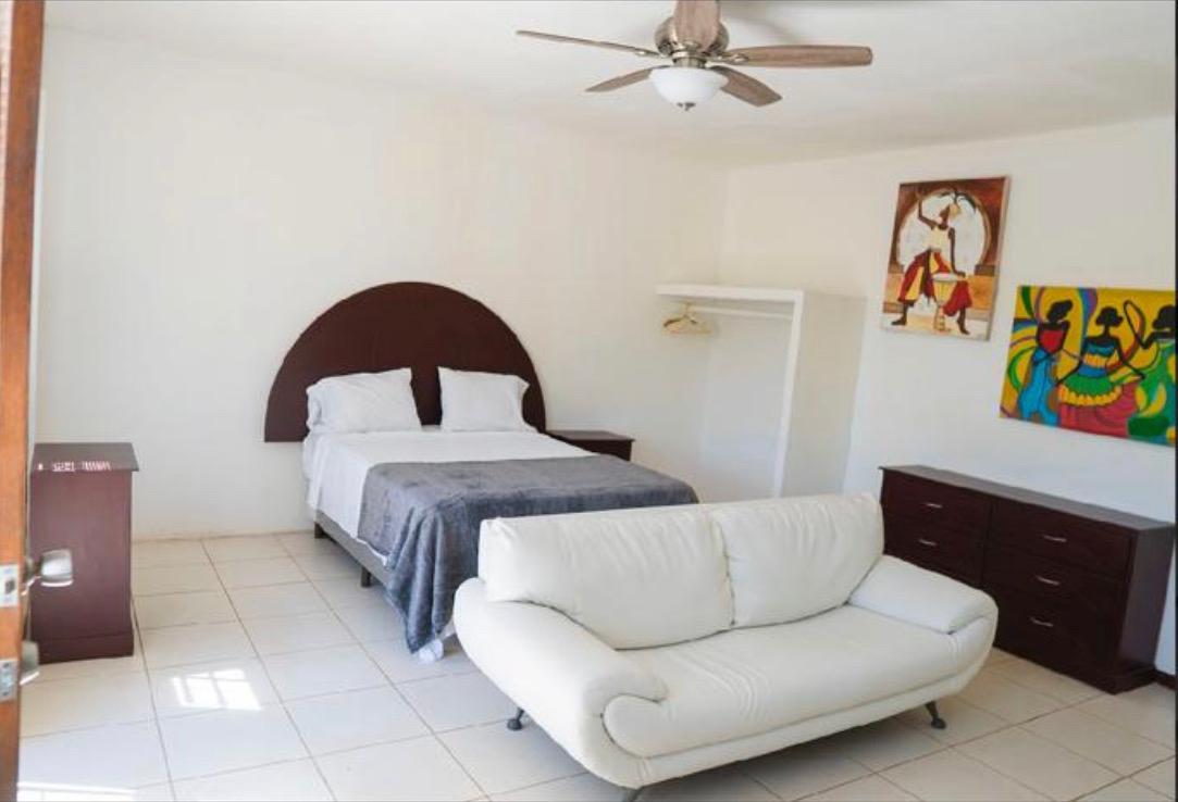 A spacious sleeping area features a large bed with white linens, a light grey throw blanket, and a ceiling fan above. A white couch is positioned near two vibrant wall art pieces, alongside a dark wood dresser and a closet with hanging space.