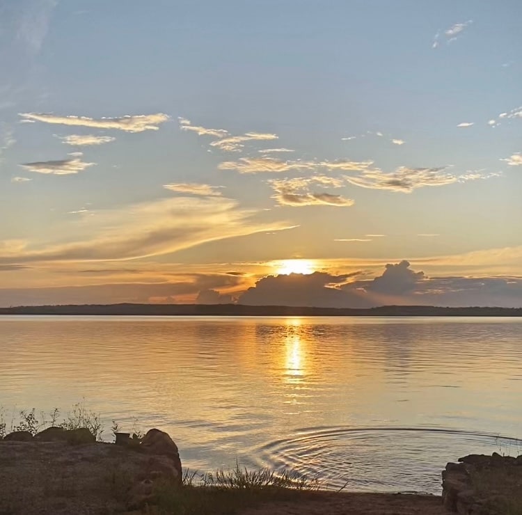 A tranquil lakeside view is presented at sunset, with soft pastel colors reflected on the water. The horizon is marked by cloud formations, and gentle ripples are visible on the lake's surface, suggesting a serene atmosphere.