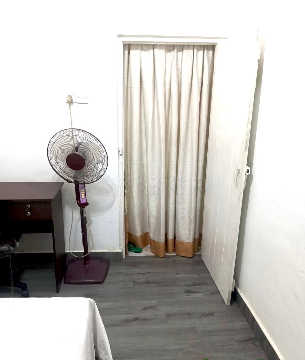 A simple room interior is depicted, featuring a desk with a chair and a standing fan. A closed door with curtains obscures another room, while light-colored walls and a light wooden floor contribute to the space's overall simplicity.