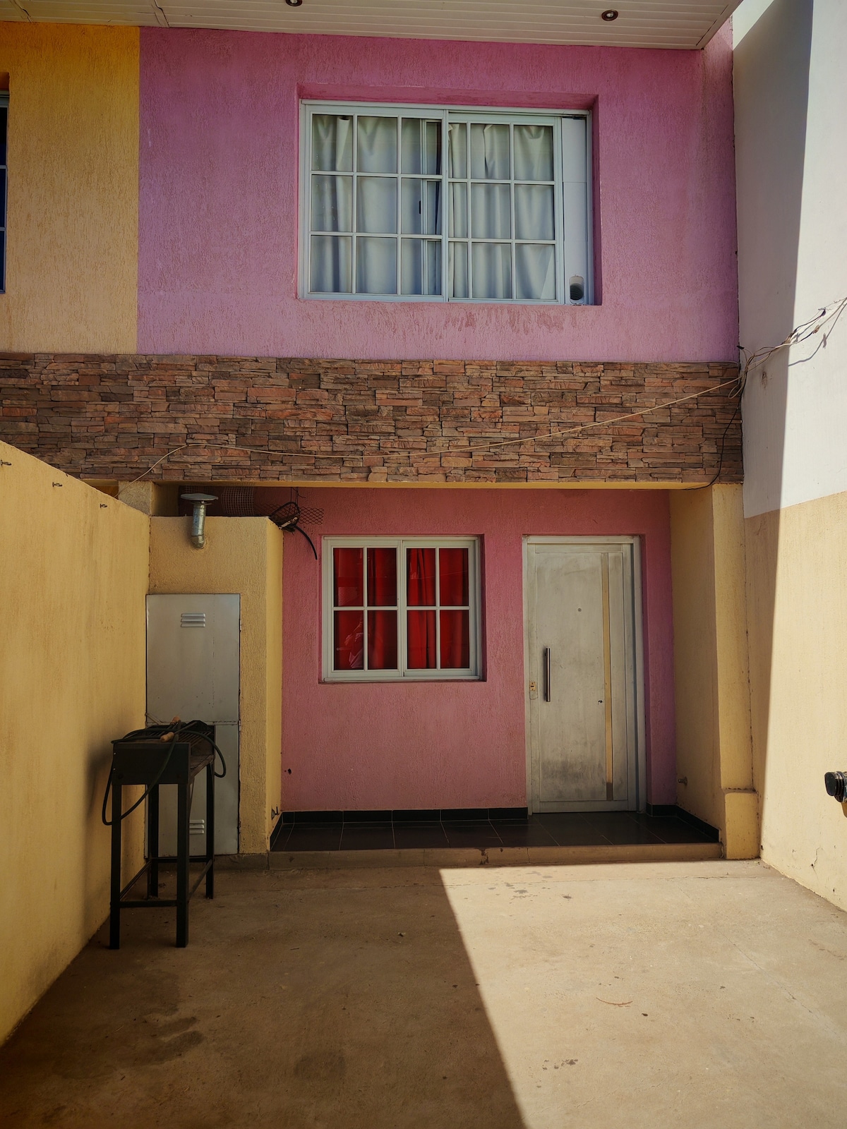 An outdoor area is visible, featuring a textured stone wall and brightly painted yellow and pink exteriors. A door stands at the center, flanked by a small window adorned with white curtains. A metal table is positioned against the wall, allowing for additional functionality.