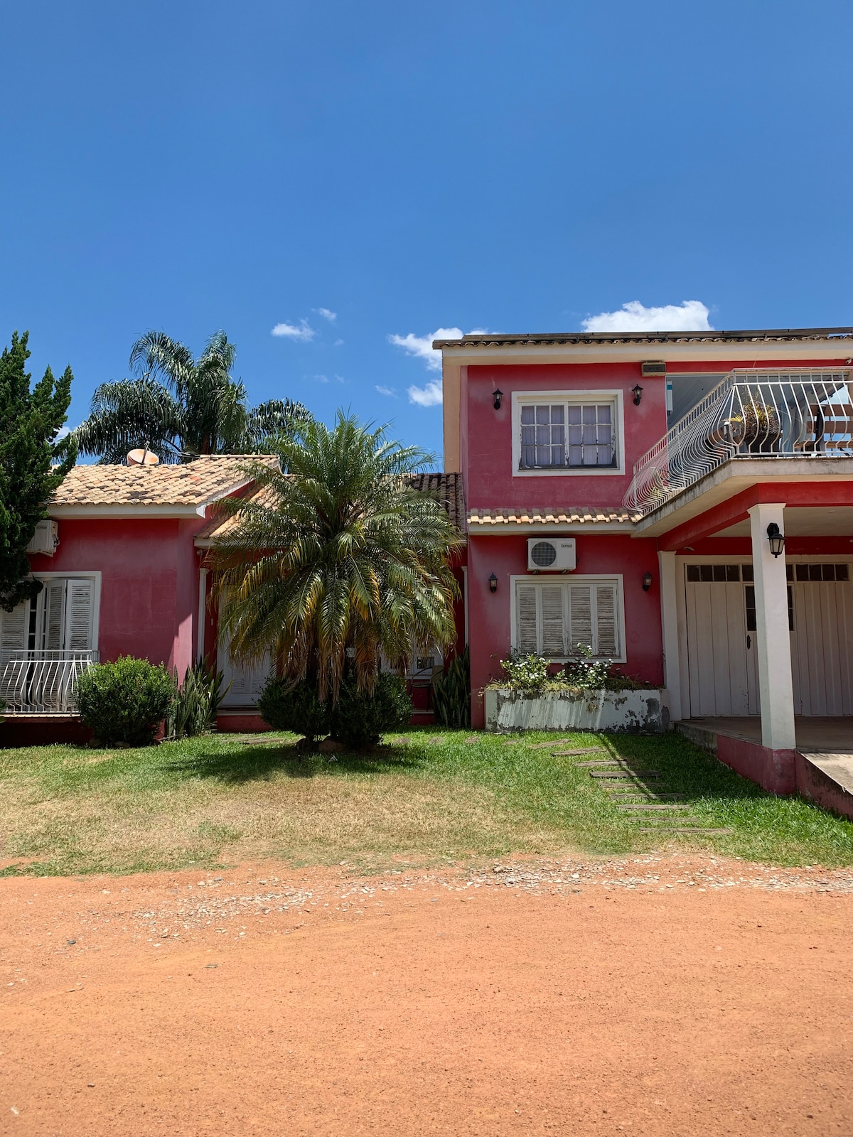 A two-story house is presented with a vibrant pink exterior, surrounded by well-maintained landscaping. A palm tree stands in the foreground, complementing the bright blue sky overhead. A balcony features floral accents, adding character to the property.
