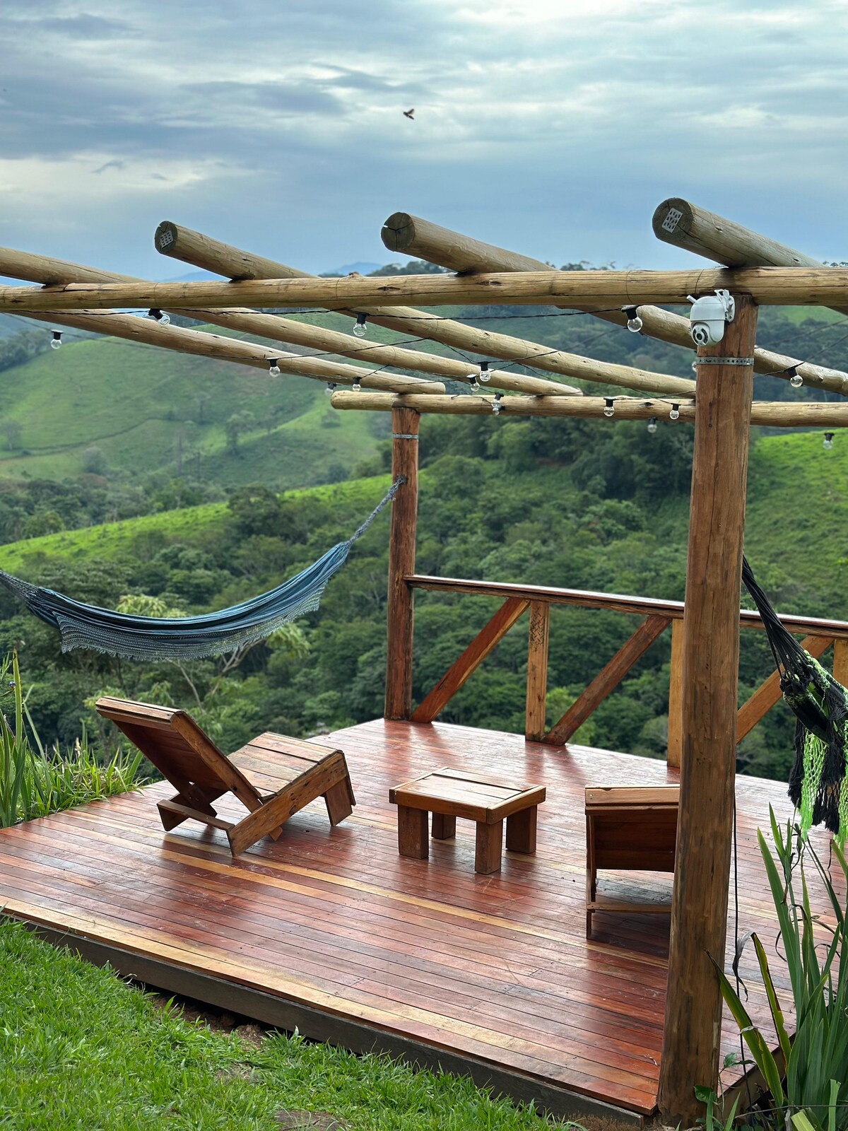 A wooden deck features two lounge chairs and a small table, surrounded by lush green hills. A hammock hangs from the structure, providing a cozy spot to relax. The area is partially covered, with string lights enhancing the natural setting.