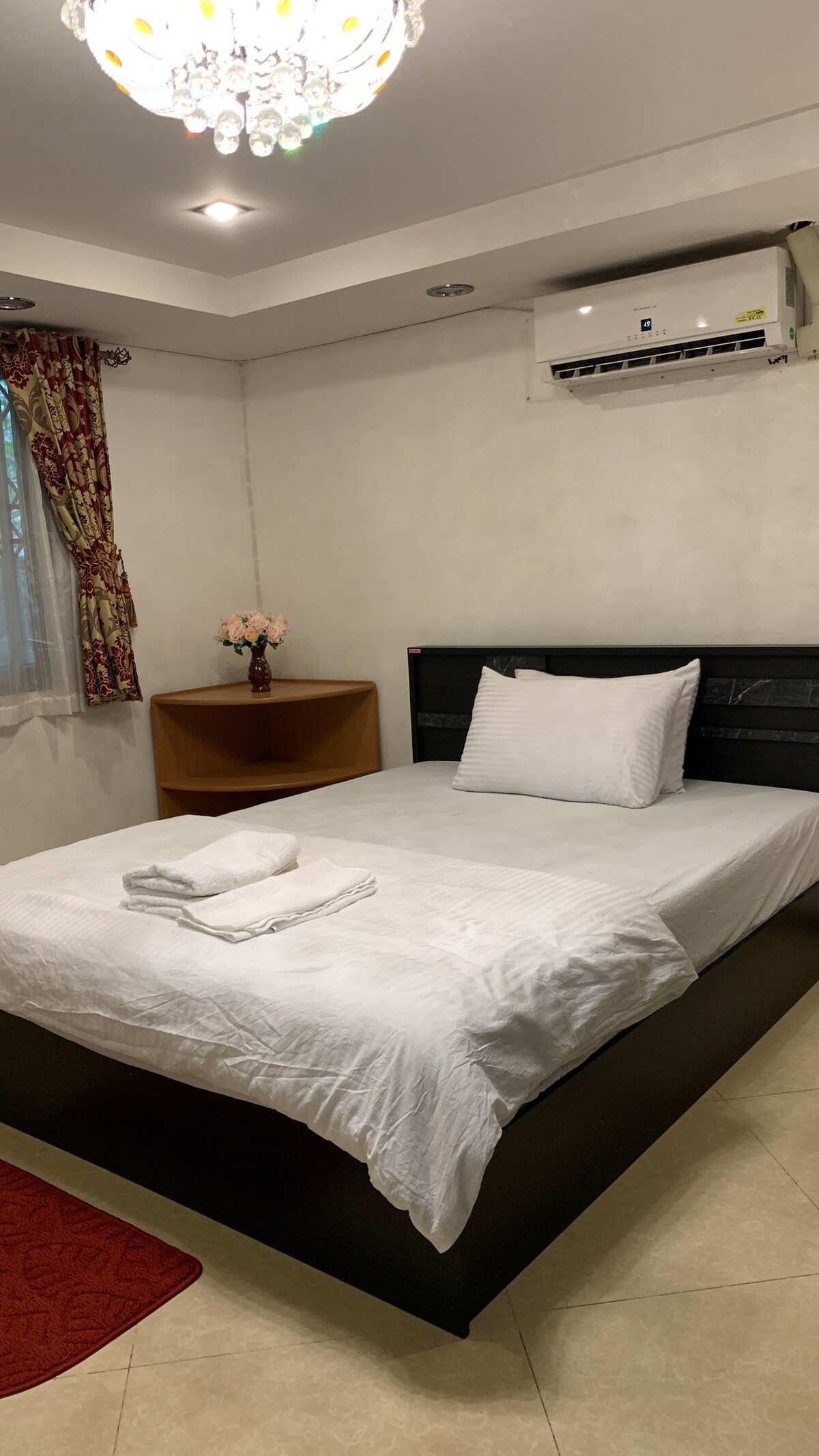 A cozy bedroom features a spacious bed adorned with white linens and two neatly arranged towels. Natural light filters through a nearby window, complemented by decorative curtains. A bedside table holds a small vase with flowers, and a subtle chandelier adds soft illumination.