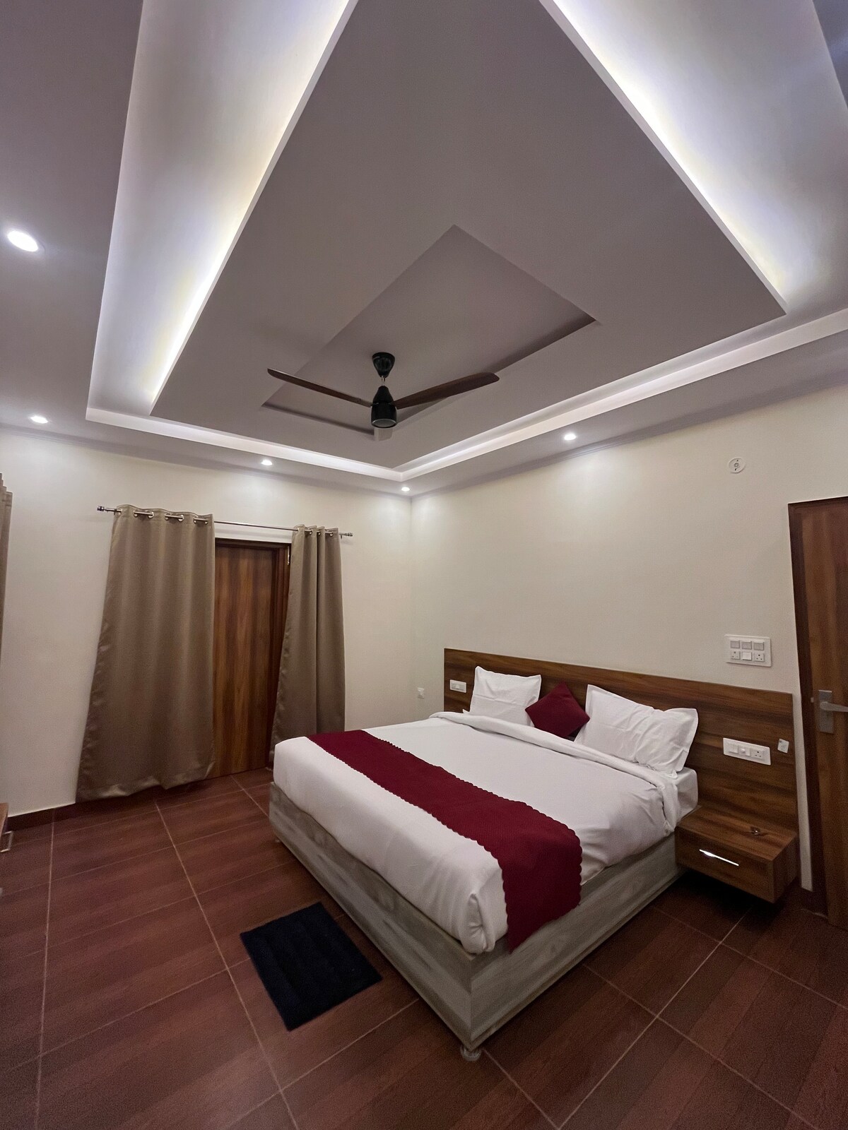 A spacious bedroom is illuminated by soft lighting, featuring a large bed with red accents. The room includes light-colored walls and elegant curtains that frame the window. A ceiling fan is centered above, contributing to a comfortable atmosphere.