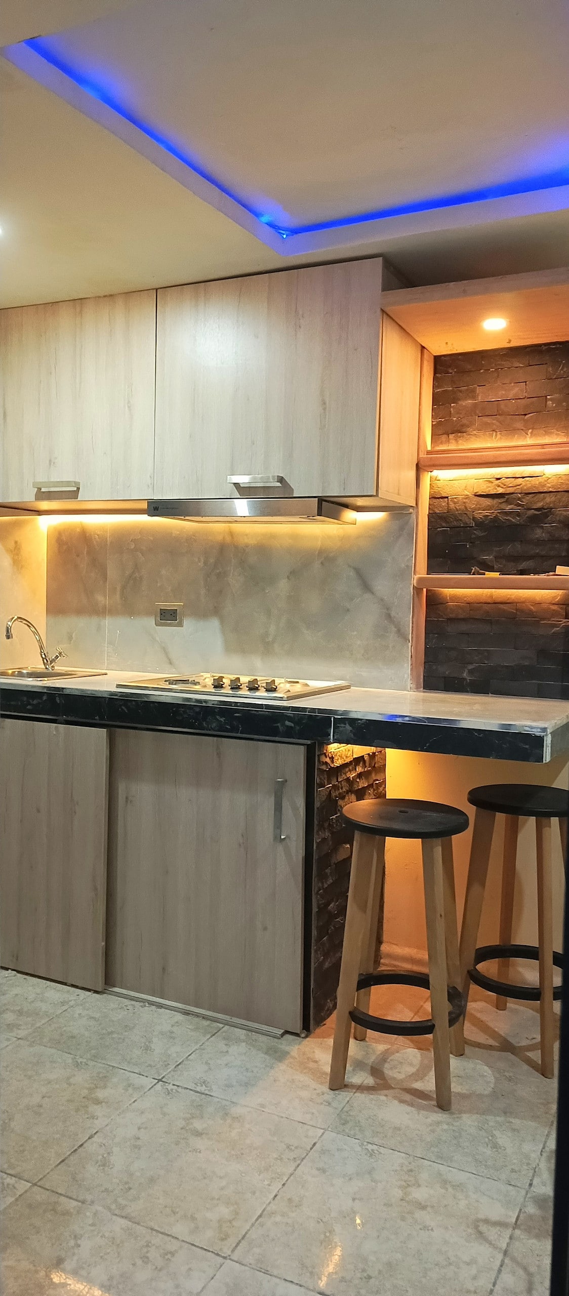 The kitchen area features wooden cabinetry with a light finish, complemented by sleek countertops. Under-cabinet lighting illuminates the workspace, while a gas stovetop is positioned against a marble-like backsplash. Two wooden stools provide seating at the countertop for casual dining.