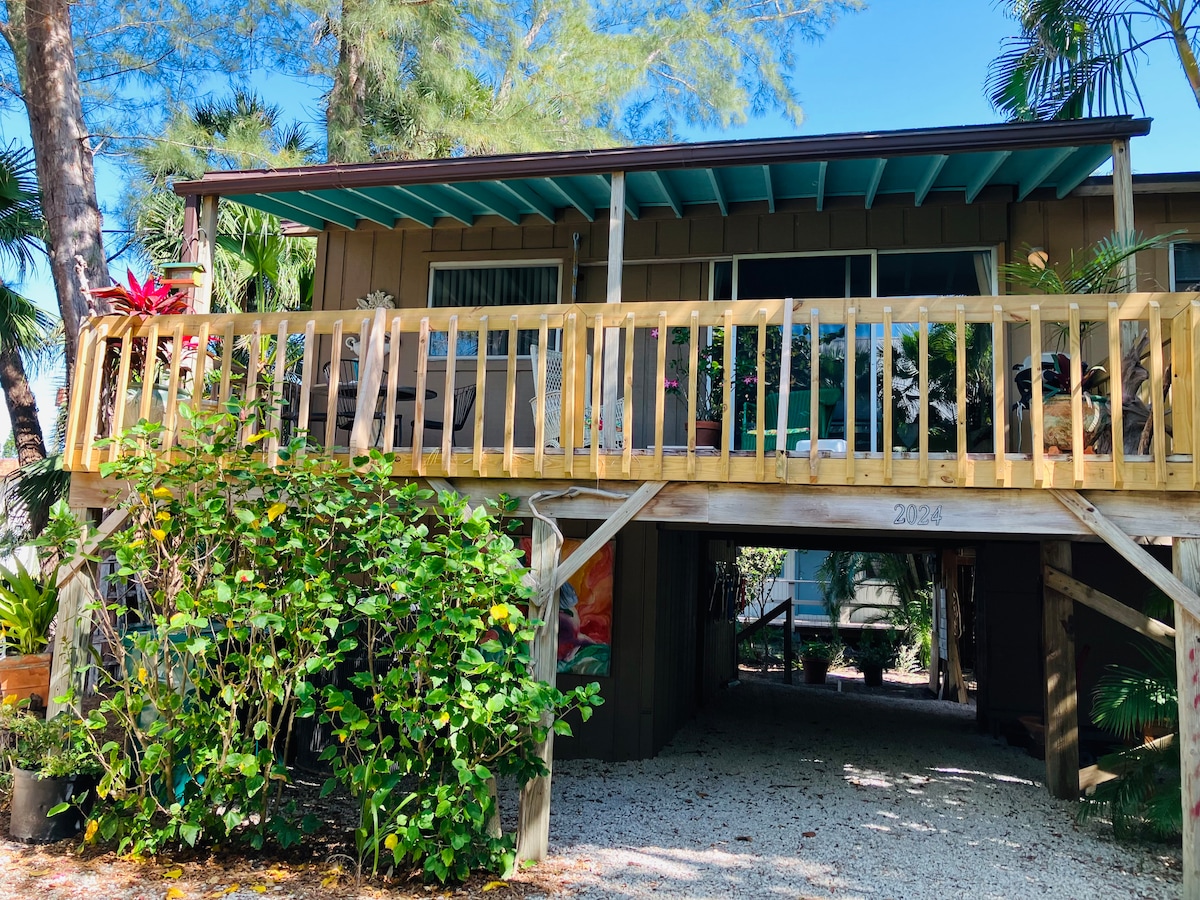 The stilt home is nestled among lush greenery, featuring a spacious wooden deck with railing overlooking the ground level. Large windows allow for natural light, while tropical plants are visible around the entrance, contributing to the inviting exterior.