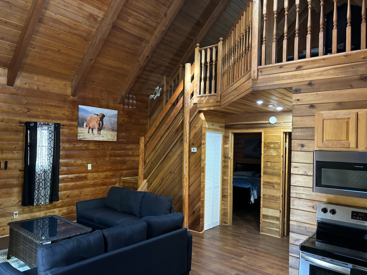 A rustic interior features exposed wood beams and a warm wooden staircase leading to a loft area. A dark sofa sits in front of a glass-top coffee table, with a mounted artwork depicting a landscape visible on the wall. Bright windows are covered by dark curtains.
