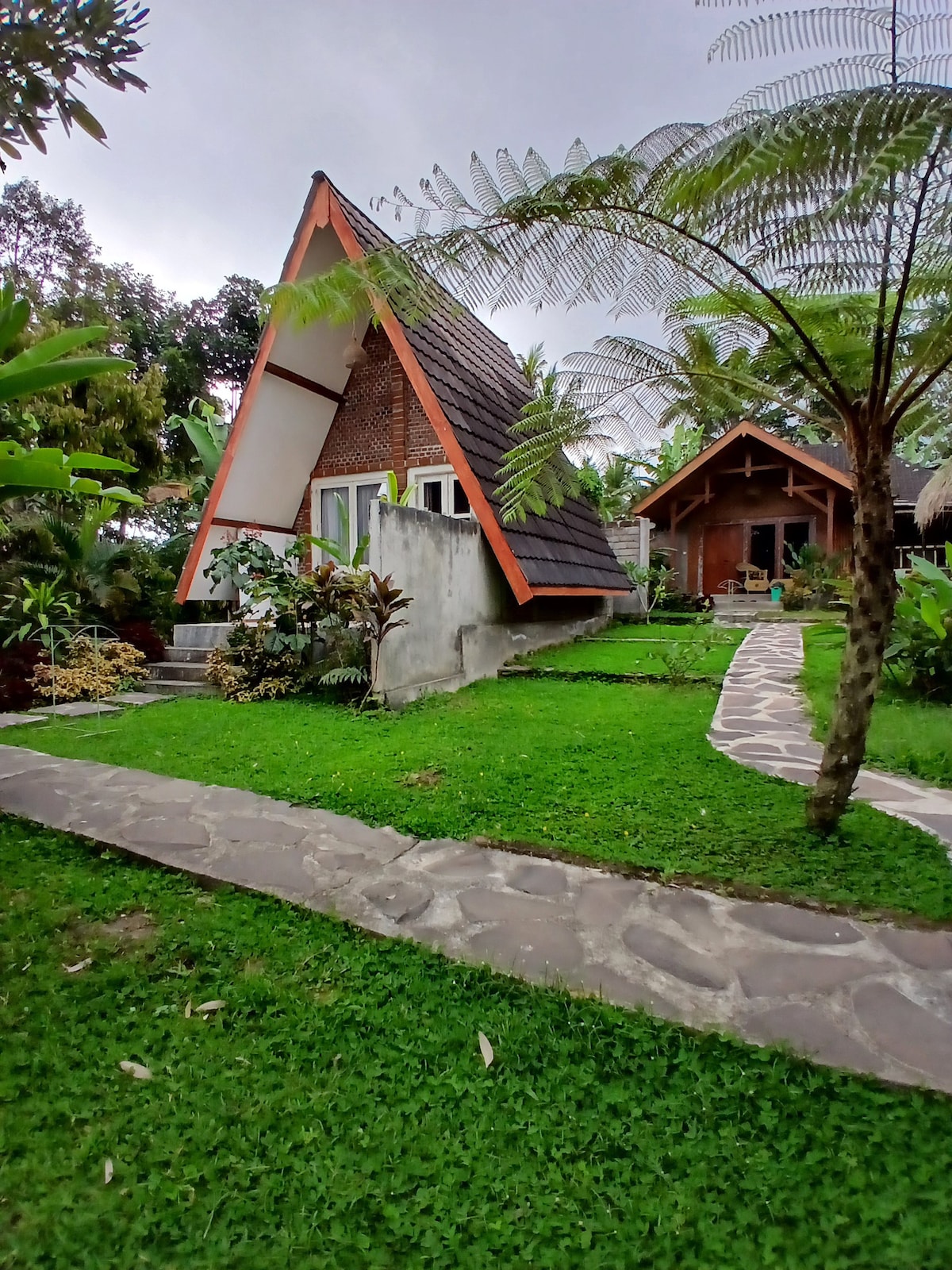 A charming two-story structure with a distinctive A-frame design is set amidst lush greenery. The surrounding landscape features well-maintained gardens, tropical plants, and a stone pathway leading to the entrance, inviting visitors to explore the peaceful surroundings.